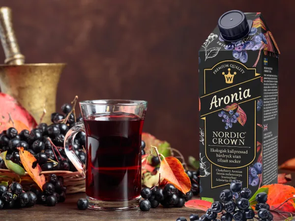 Glas med mörkröd aroniadryck bredvid en kartong, höstlöv och bär på bordet