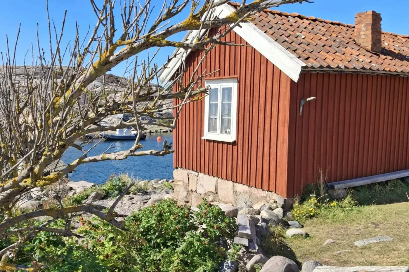 Röd sjöbod vid vattnet i Bohuslän, klippor och båtar i bakgrunden